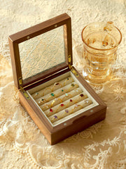 A wooden box with a glass lid displays CAMELLIA's Mystery Gemstone Rings on a lace tablecloth, next to a transparent glass cup of light-colored liquid. Glimpses of yellow gold and sterling silver add elegance to the assorted colorful rings.