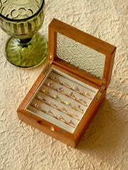 A CAMELLIA Mystery Gemstone Ring rests in a wooden box with an ornate glass lid among vibrant rings, some in sterling silver. A green glass goblet is nearby on a textured surface.
