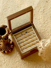 A wooden jewelry box with a glass lid showcases an assortment of jewelry, including the CAMELLIA Mystery Gemstone Ring in sterling silver. Nearby, a vintage candlestick holder and lace fabric rest on the textured surface, completing an elegant display.