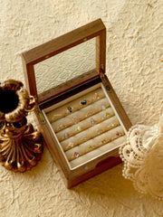 A wooden jewelry box with a glass lid showcases an assortment of jewelry, including the CAMELLIA Mystery Gemstone Ring in sterling silver. Nearby, a vintage candlestick holder and lace fabric rest on the textured surface, completing an elegant display.