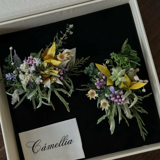 Two intricate floral brooches with green, yellow, purple, and white artificial flowers and leaves, arranged on a black velvet base inside a box labeled