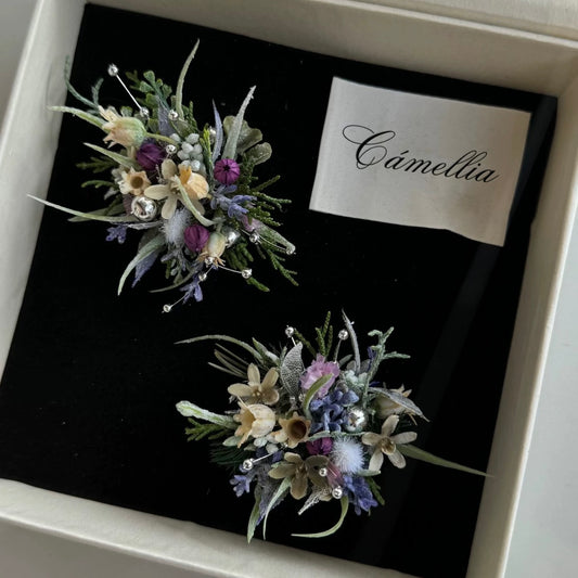 Two floral brooches with purple, white, and green artificial flowers are displayed on black velvet in a box labeled
