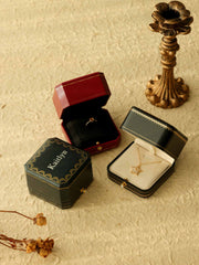 Two Customized Vintage Jewelry Boxes by CAMELLIA: one red containing a ring and one green with a pendant, complemented by an ornate candlestick and dried flowers in the background.
