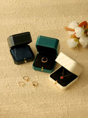 On a textured surface, three CAMELLIA "Customized Vintage Jewelry Boxes": one open with a ring, one holding a necklace, and an empty one. Nearby are two more rings, with white flowers in the background.