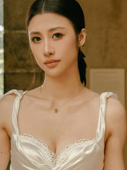 A woman with long dark hair tied back, wearing a white lace-trimmed top and the delicate New York Peridot Pentagram Necklace by CAMELLIA, stands indoors.