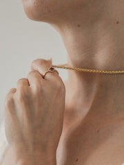 A person holds a 14K gold chain necklace near their neck with one hand while wearing the CAMELLIA 14K Solid Gold Carena Garnet Ring, featuring a vintage-inspired design and round garnet stone, on their finger.