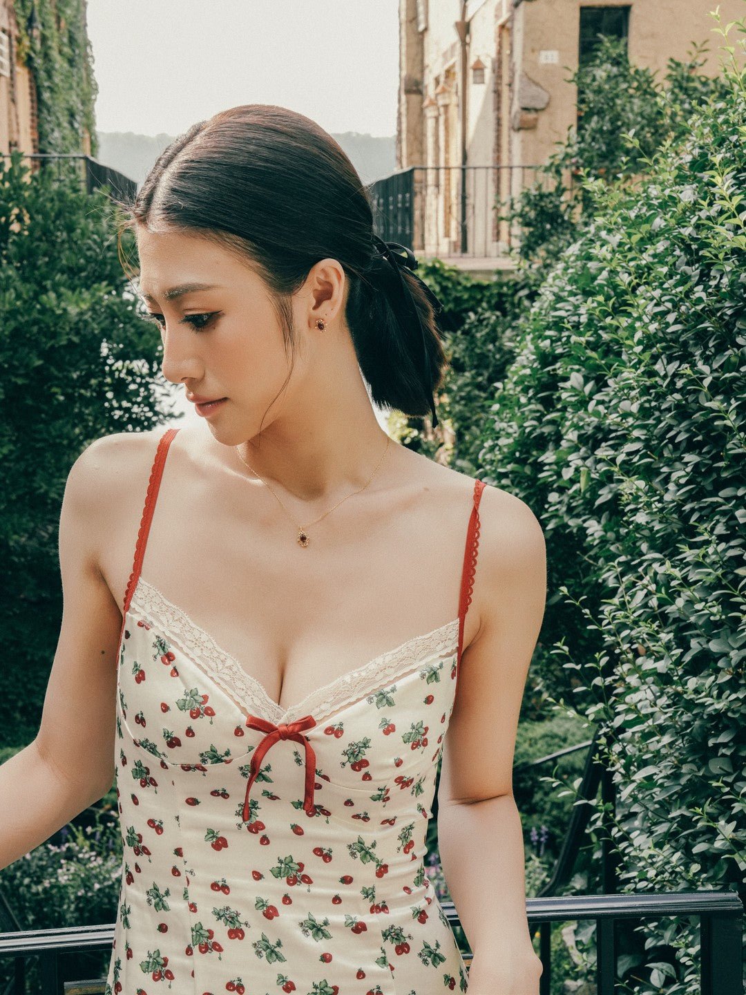 Wearing a floral dress with red straps, a person stands outdoors amidst lush greenery and buildings. Elegant Vivien Garnet earrings from CAMELLIA gleam in the sunlight, adding sophistication to the vibrant scene.