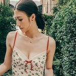 Wearing a floral dress with red straps, a person stands outdoors amidst lush greenery and buildings. Elegant Vivien Garnet earrings from CAMELLIA gleam in the sunlight, adding sophistication to the vibrant scene.
