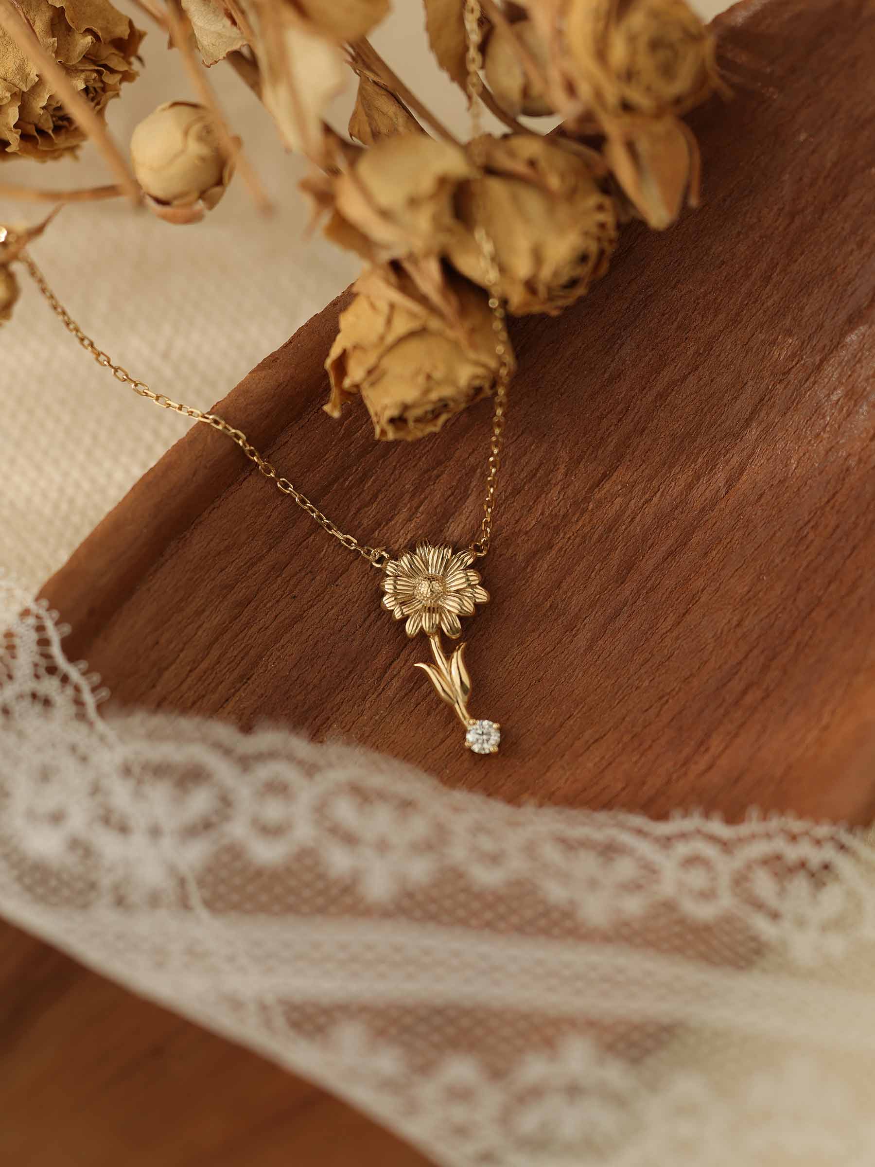 The Nerissa Daisy Diamond Necklace from CAMELLIA, made of 14K Yellow Gold Vermeil and hypoallergenic for sensitive skin, features a floral pendant with diamond details. It's beautifully showcased on a wooden board with dried flowers and lace fabric.