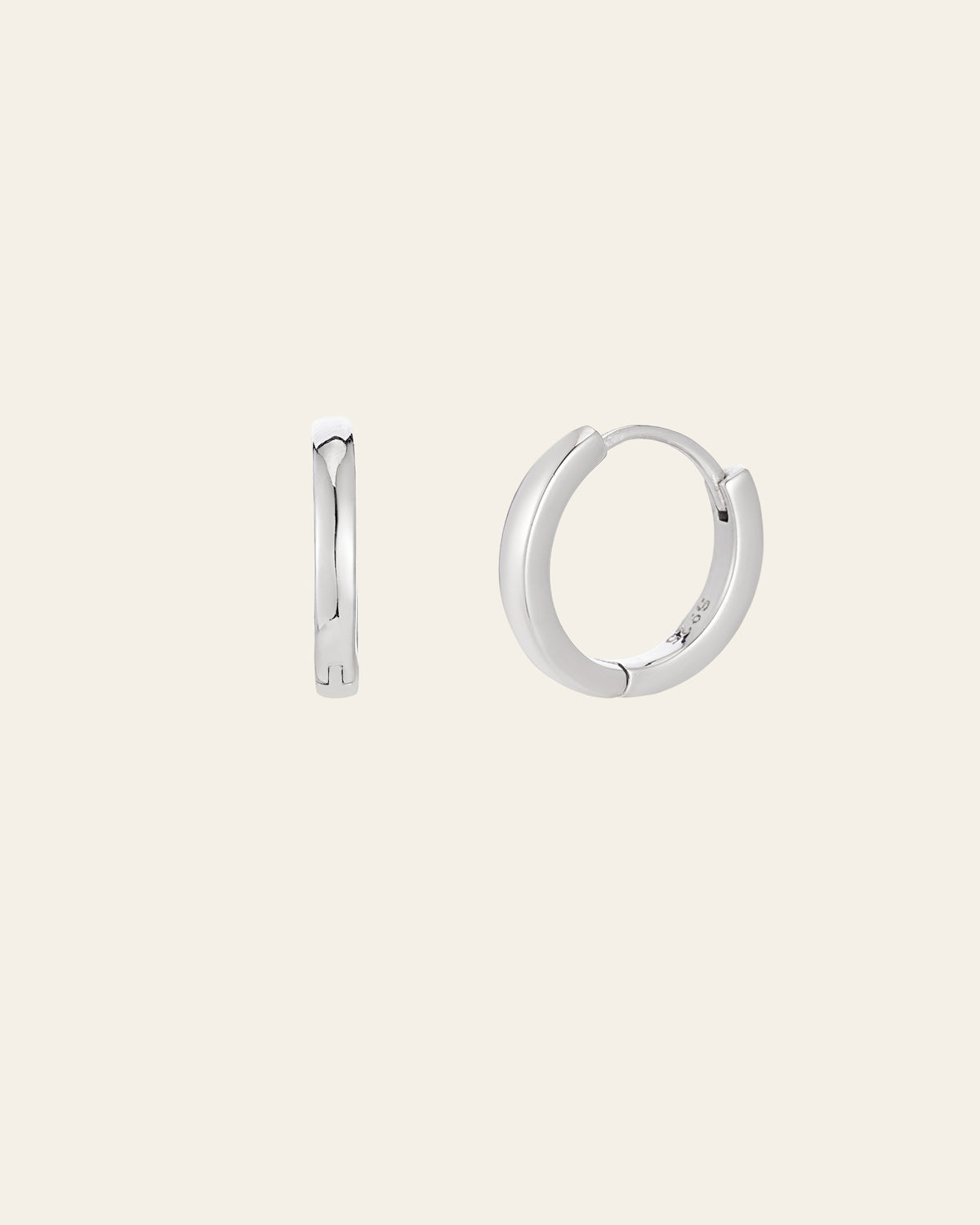 Displayed against a plain beige background, the 15mm Mother Hoops by CAMELLIA showcase their smooth, simple design. One earring is shown from the side and the other from the front, highlighting their elegance. Crafted in sterling silver to be hypoallergenic, they offer everyday comfort.