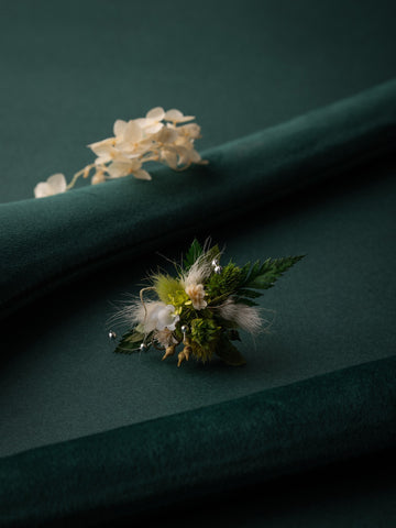 A handcrafted floral arrangement with green and white blossoms rests on a dark green fabric, complemented by CAMELLIA's Verdant Dreams Flower Ring featuring another white bloom nearby.