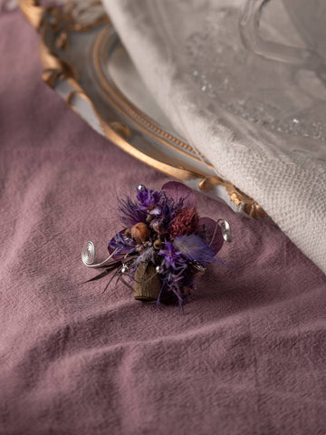 The Amethyst Glow Flower Ring by CAMELLIA evokes handcrafted elegance with its small, intricate purple and brown design, reminiscent of a boutonniere on mauve fabric near a decorative plate edge and white cloth. Made with care in New York, it’s truly an adjustable masterpiece.