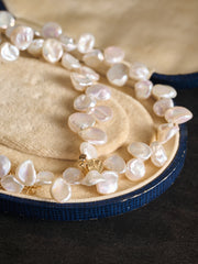 The CAMELLIA Petal Pearl Necklace, featuring irregular freshwater pearls and a gold clasp, is displayed on a beige circular jewelry case with navy blue trim.