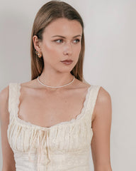 A woman with long hair, dressed in a lace-trimmed cream dress and 14K Yellow Gold Vermeil hoop earrings, wears a CAMELLIA 3mm Pearl Chain Necklace while looking to the side.