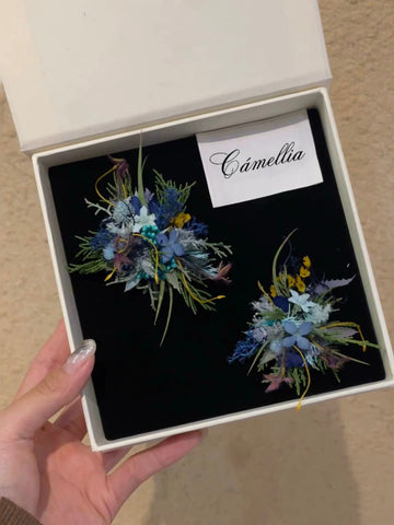 A person holds an open box showing two handcrafted pins on a black background, with a card marked "CAMELLIA" alongside the Customized Unique Flower Ring.