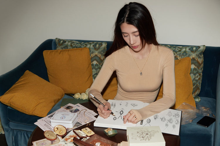 A woman sits on a couch, sketching jewelry designs in a notebook on a table with various papers, snacks, and a box in front of her.