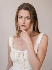 A woman with long light brown hair in a sleeveless white dress gazes at the camera, her hand near her chin showcasing the CAMELLIA Evermine Moissanite Band with pavé-set accents, set against a neutral background.