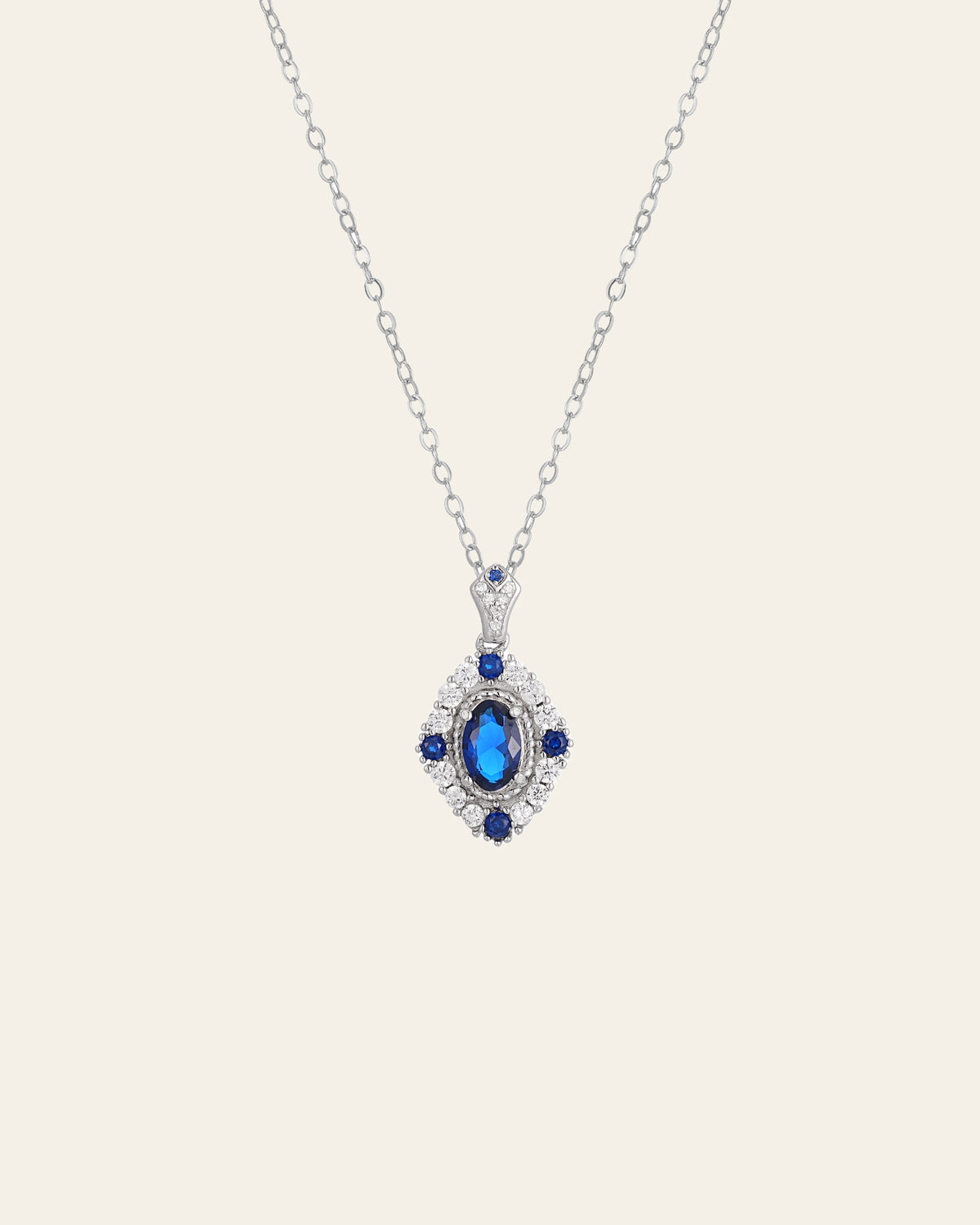 The Eta Sapphire Necklace by CAMELLIA is a sterling silver piece featuring a central sapphire pendant, surrounded by smaller blue stones and clear embellishments.