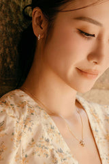 A woman with long hair and a floral dress, accessorized with the elegant "Quillie Cz Hoops" from CAMELLIA in 14K Yellow Gold Vermeil, gazes down with a soft smile.
