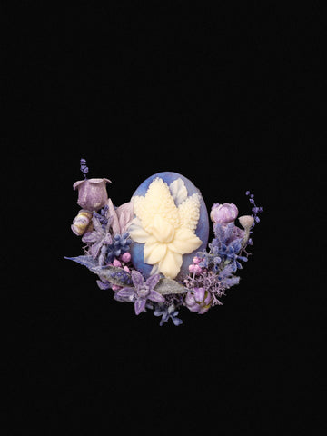 A detailed carved white flower sits at the center of a blue egg, framed by purple and pink floral accents on a black backdrop. This exquisite design embodies CAMELLIA's Hyacinthus Birth Flower Ring, handcrafted in New York.