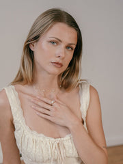 The woman with long blonde hair, wearing a light-colored lace-detailed top and the CAMELLIA Etheral Moissanite Band, gazes at the camera. Her hand resting on her chest and a neutral expression exude understated elegance.