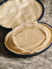 The CAMELLIA Classic Pearl Strand Necklace with a gold clasp is showcased in an open, oval velvet jewelry box on a textured surface.