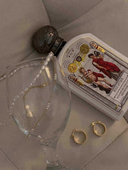 A CAMELLIA Polly Pearl Chain Necklace and a 14K Yellow Gold Vermeil chain embellish a wine glass tipped on a couch, alongside a decorated bottle and two hypoallergenic sterling silver hoop earrings.