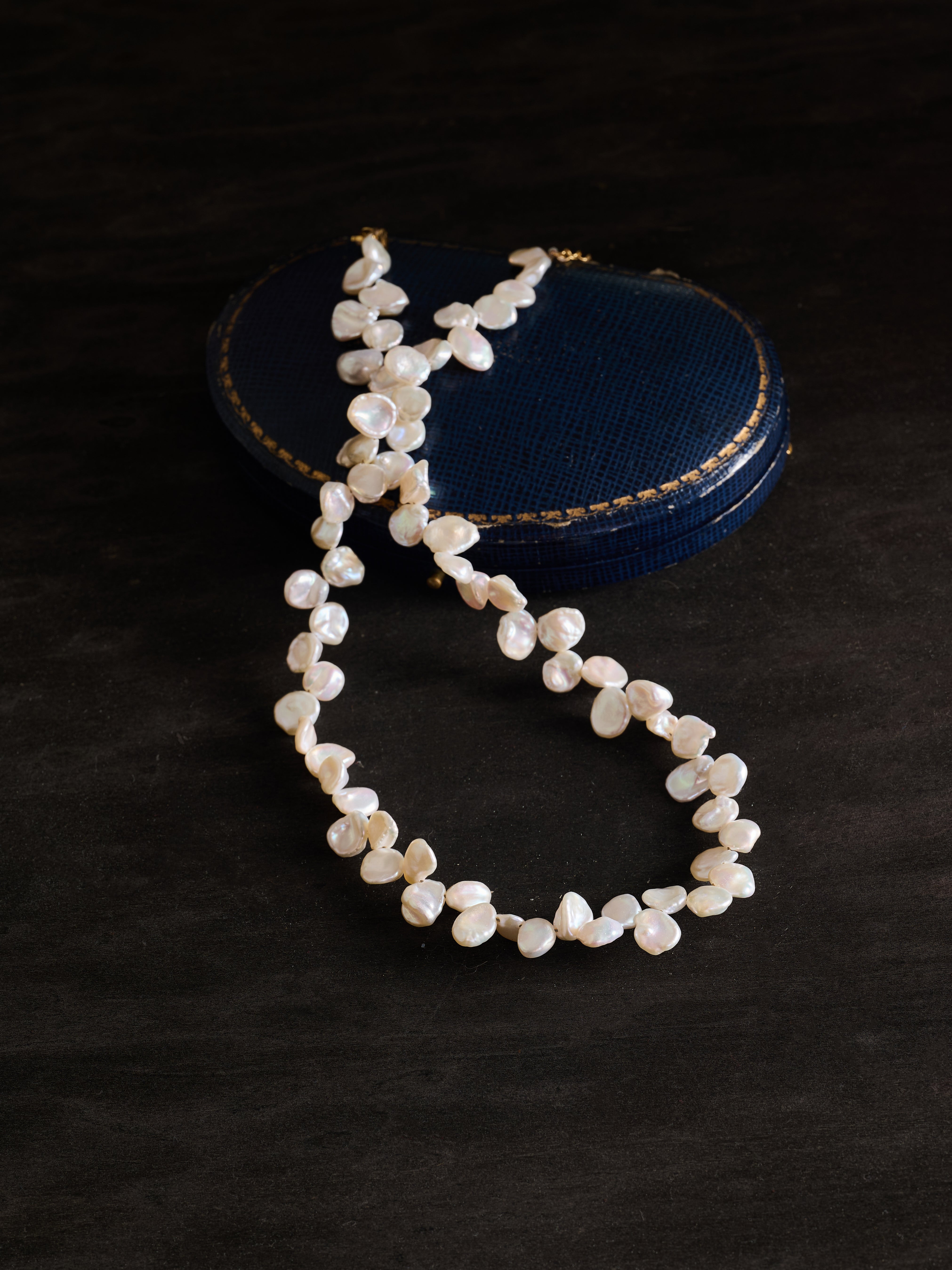 The CAMELLIA Petal Pearl Necklace is shown on a dark surface, with a round, textured dark blue case in the background.