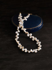 The CAMELLIA Petal Pearl Necklace is shown on a dark surface, with a round, textured dark blue case in the background.