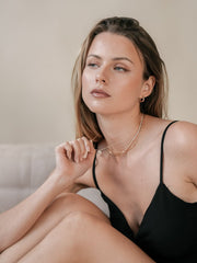 A woman with long hair sits on a couch wearing a black strappy top. With a neutral expression, she touches her layered New York Opal Pentagram Necklace by CAMELLIA, which glimmers subtly in 14K Yellow Gold Vermeil, adding an elegant touch to her look.