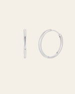 The 25mm Mother Hoops, elegant sterling silver earrings, are showcased against a light backdrop. One earring is shown side-on while the other faces forward. These shiny, circular pieces are hypoallergenic for comfort and promise timeless style in any collection.