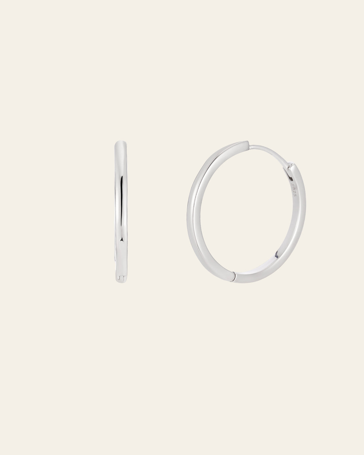 The 25mm Mother Hoops, elegant sterling silver earrings, are showcased against a light backdrop. One earring is shown side-on while the other faces forward. These shiny, circular pieces are hypoallergenic for comfort and promise timeless style in any collection.