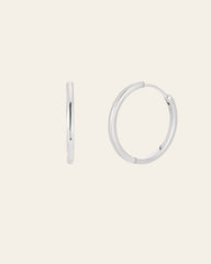 The 25mm Mother Hoops, elegant sterling silver earrings, are showcased against a light backdrop. One earring is shown side-on while the other faces forward. These shiny, circular pieces are hypoallergenic for comfort and promise timeless style in any collection.