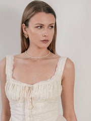 A woman with long hair wears a sleeveless, lace-trimmed white top, pearl necklace, and CAMELLIA's Infinite Hoops. She gazes sideways against a plain background, exuding elegance and hypoallergenic charm.