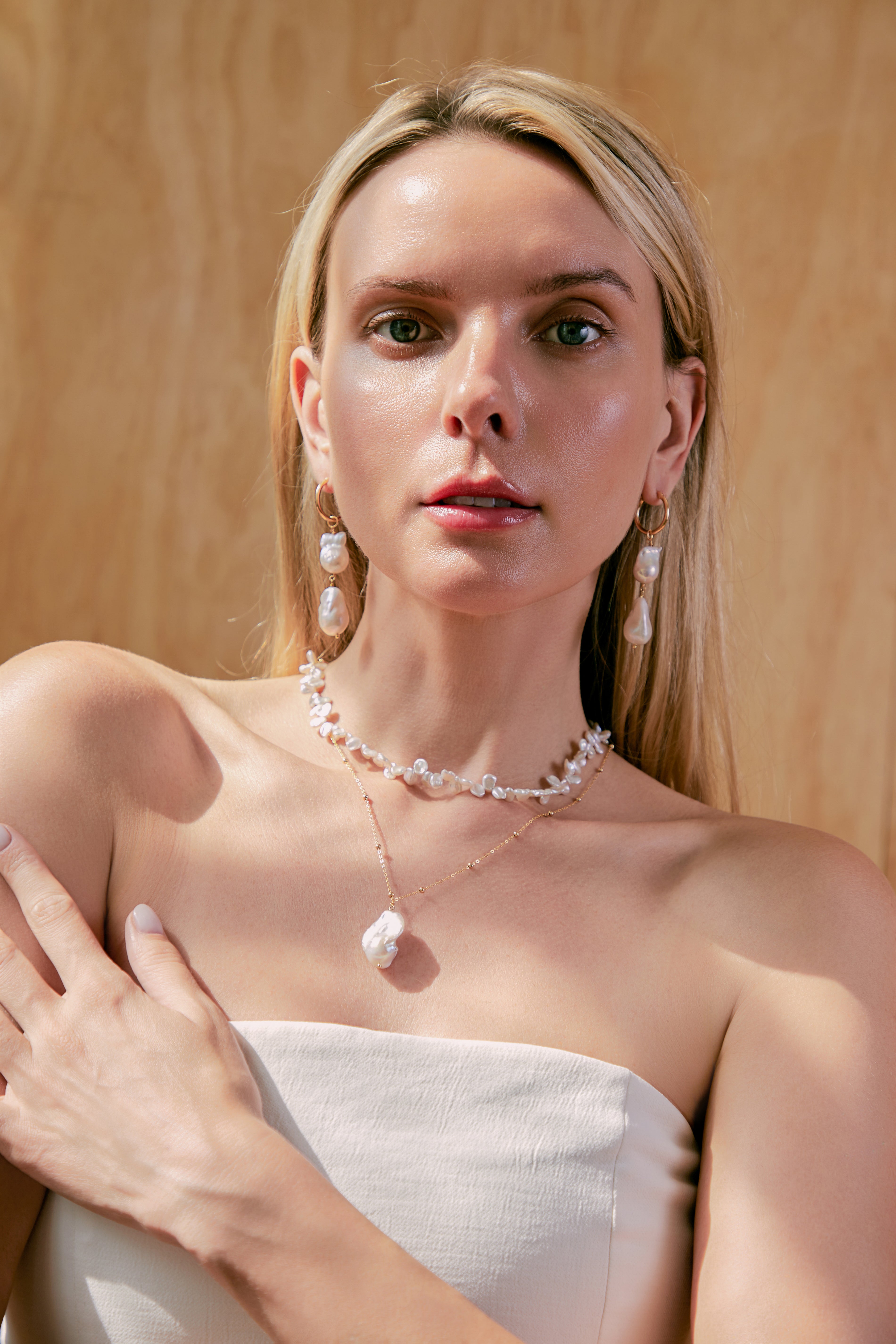 A woman with long blonde hair wears a white strapless top, pearl earrings, and the CAMELLIA Petal Pearl Necklace, standing against a light wood background.