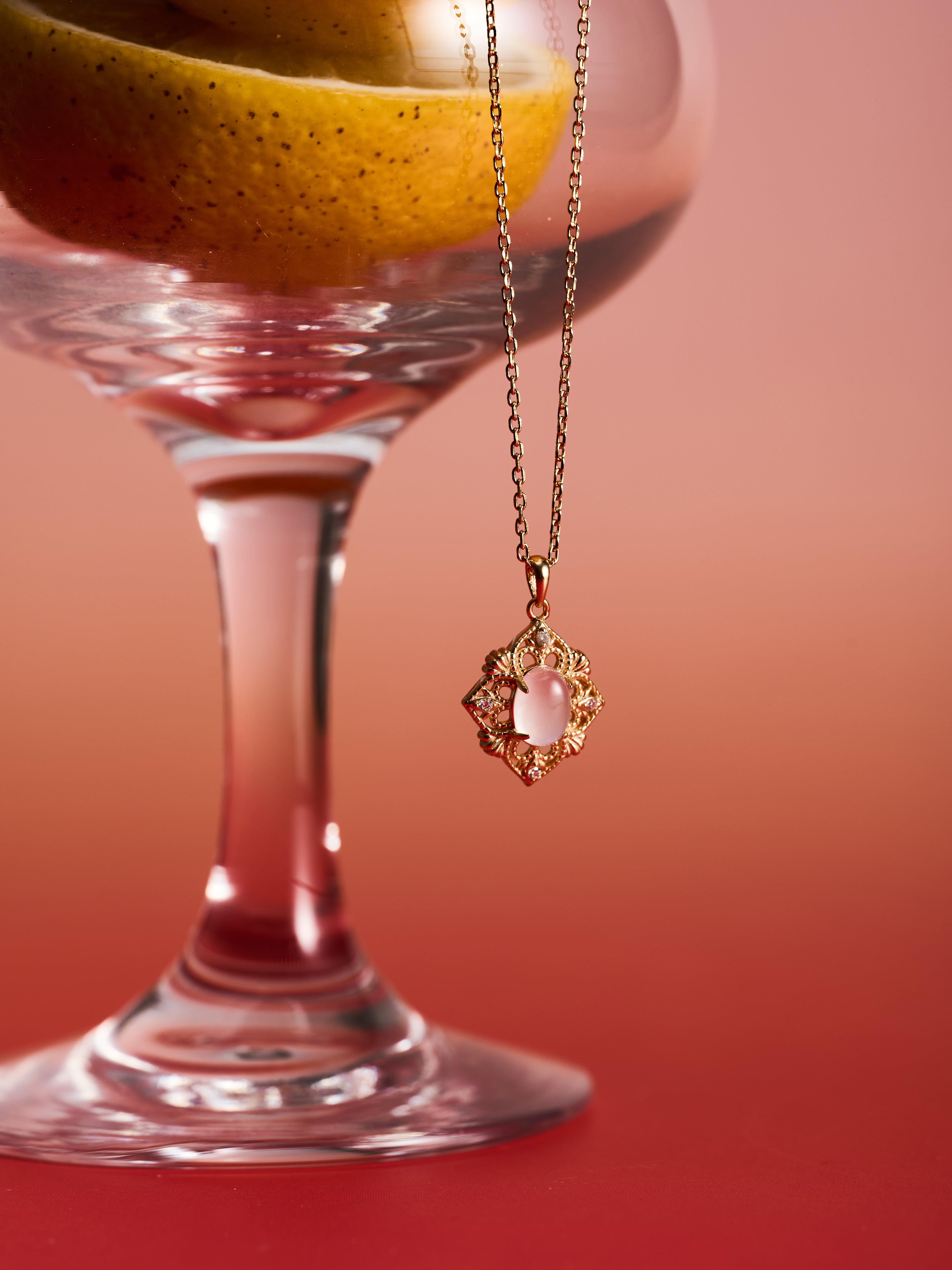 The CAMELLIA Capri Moonstone Necklace, featuring a gold vermeil chain and moonstone pendant, hangs from the rim of a glass with a pear, set against a pink gradient—ideal for vintage jewelry enthusiasts.