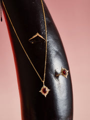 The CAMELLIA Claire Garnet Diamond Jewelry Set features a gold necklace with a garnet pendant, a matching ring, and a gold ring with clear stones, elegantly displayed on black against a pink background.