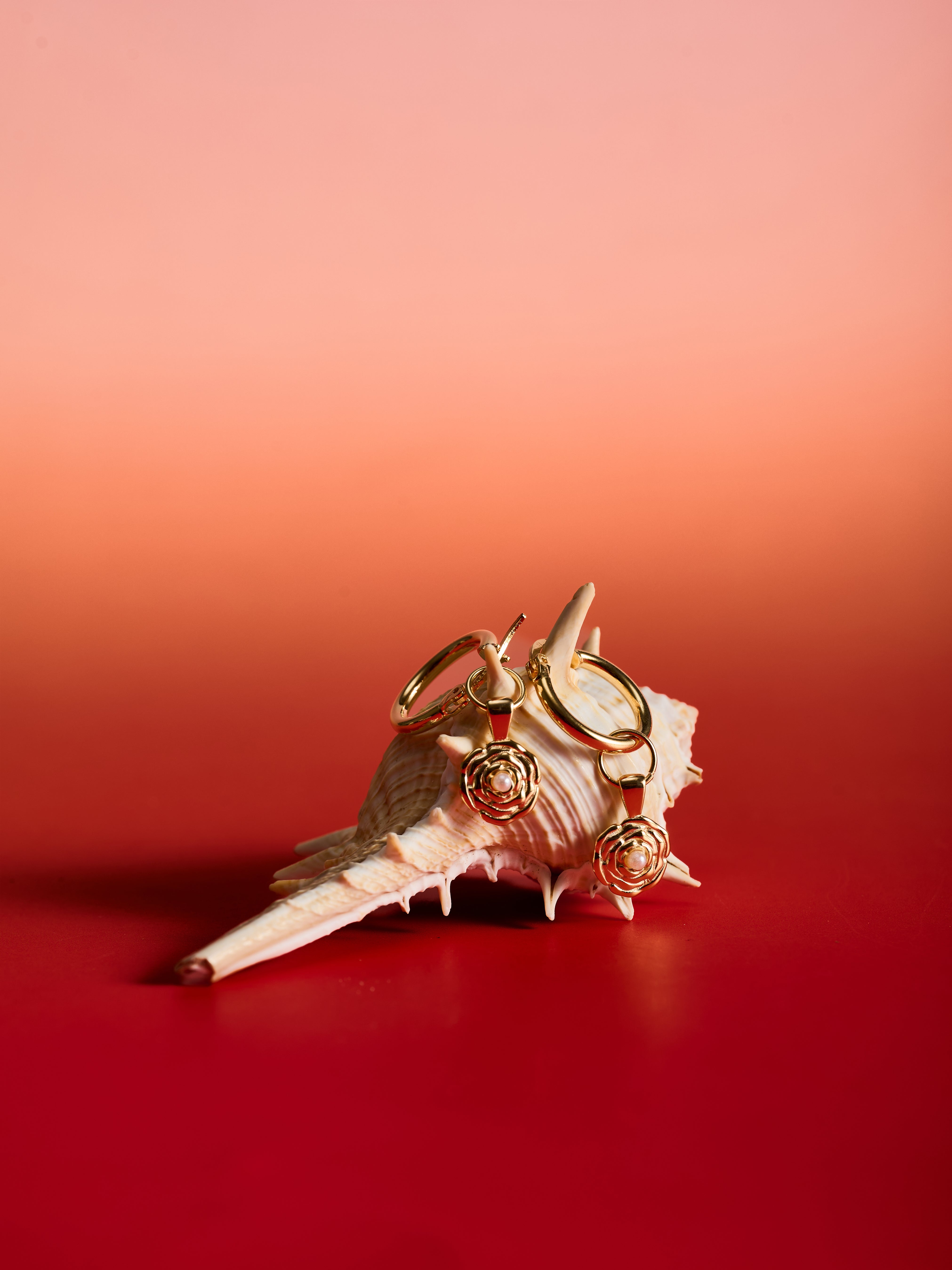 The CAMELLIA Elsa Birth Month Earrings are 14K gold vermeil earrings with rose-shaped pendants, inspired by vintage floral designs and shown on a seashell with a red gradient background.