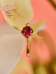 Close-up of the CAMELLIA Carena Ruby Ring, featuring a vintage-inspired red gemstone and a small clear stone, set against a softly blurred peach and cream background.