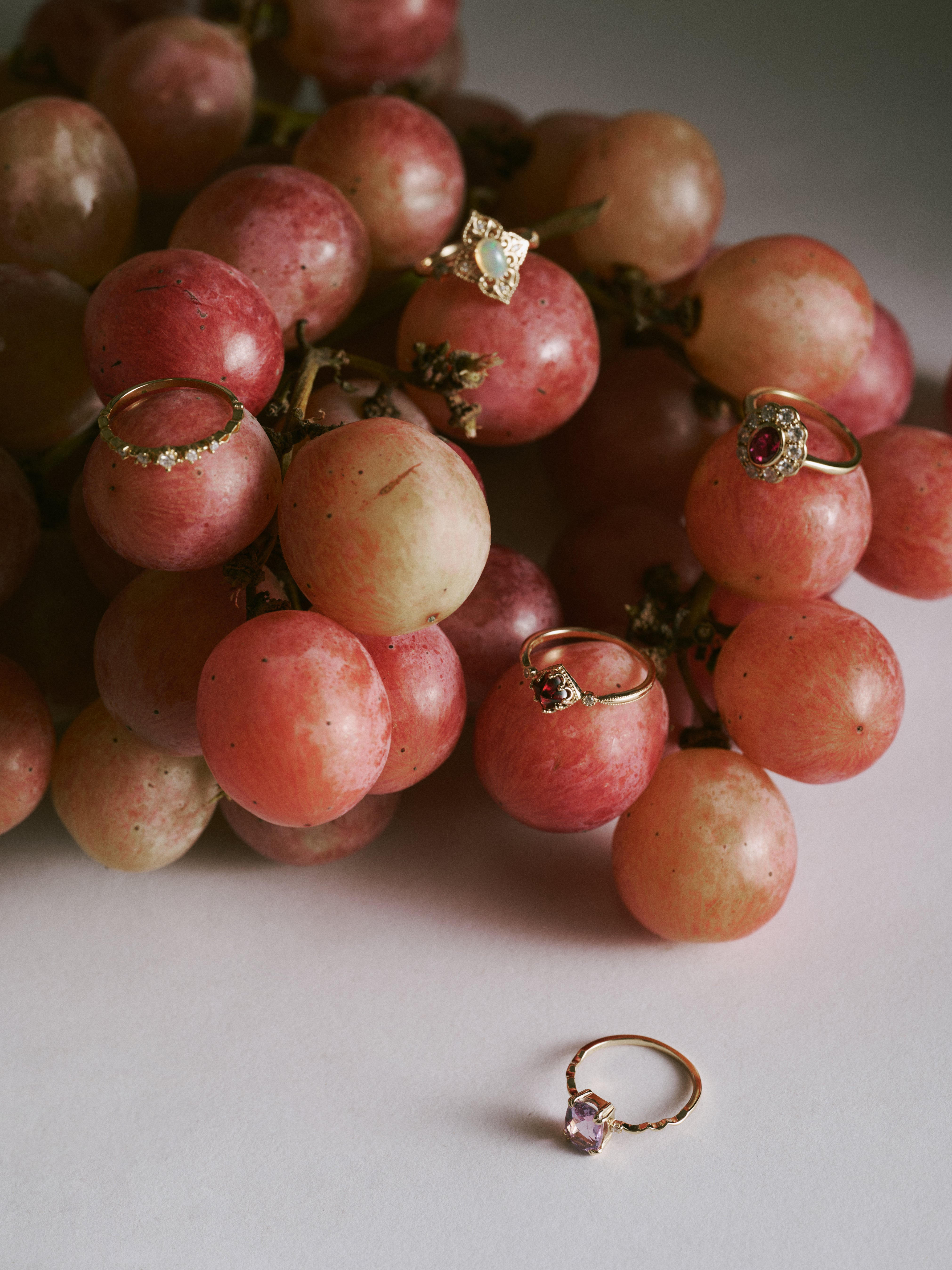 Five CAMELLIA Lunar Amethyst Rings, each featuring an amethyst gemstone, are displayed on and around a bunch of red grapes against a plain background.