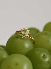 The CAMELLIA Aurora Peridot Ring, featuring a green center stone and sparkling accents, is elegantly displayed on a cluster of green grapes.