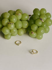 Two clusters of green grapes are decorated with several gold rings, including the CAMELLIA Eden Peridot Ring and a vintage-inspired design, some set with stones, elegantly displayed on and around the grapes against a plain background.