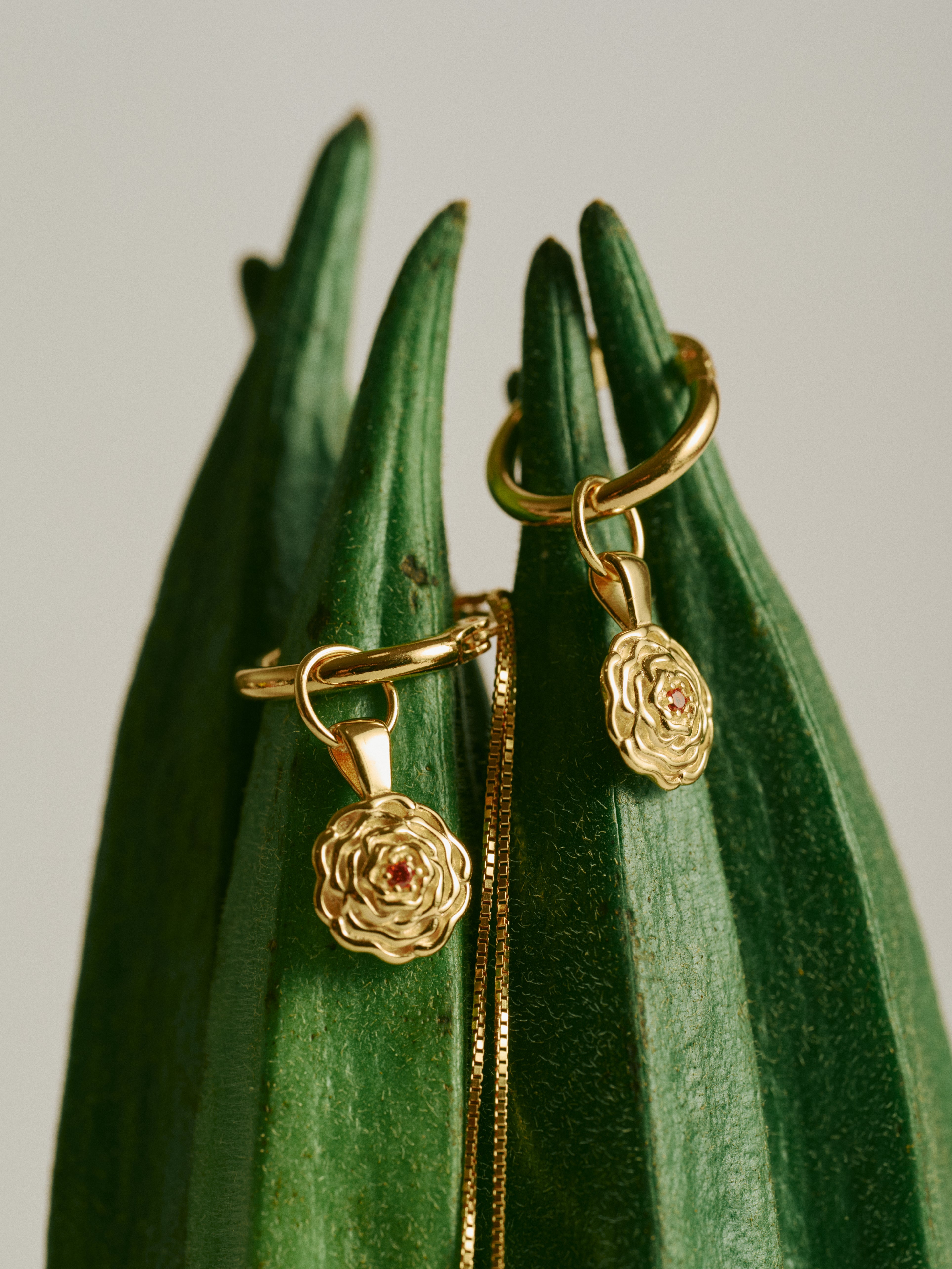 The CAMELLIA Elsa Birth Month Earrings, featuring a gold vintage floral design with small red stones, hang on green plant leaves with a gold chain draped between them.