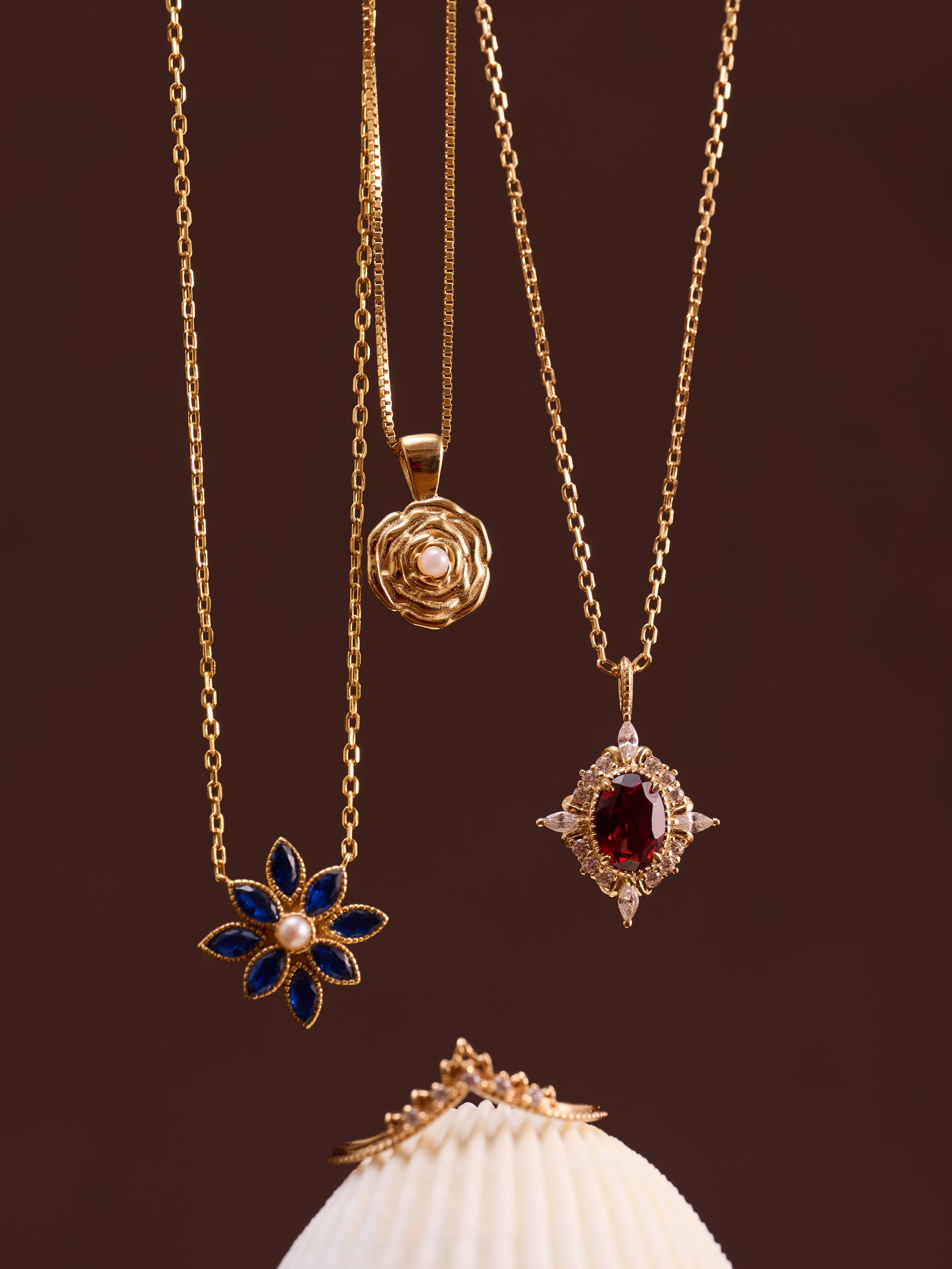 The CAMELLIA Claire Garnet Diamond Necklace hangs above gold rings on a white seashell against a brown background, highlighting elegant birthstone jewelry with a unique pendant.