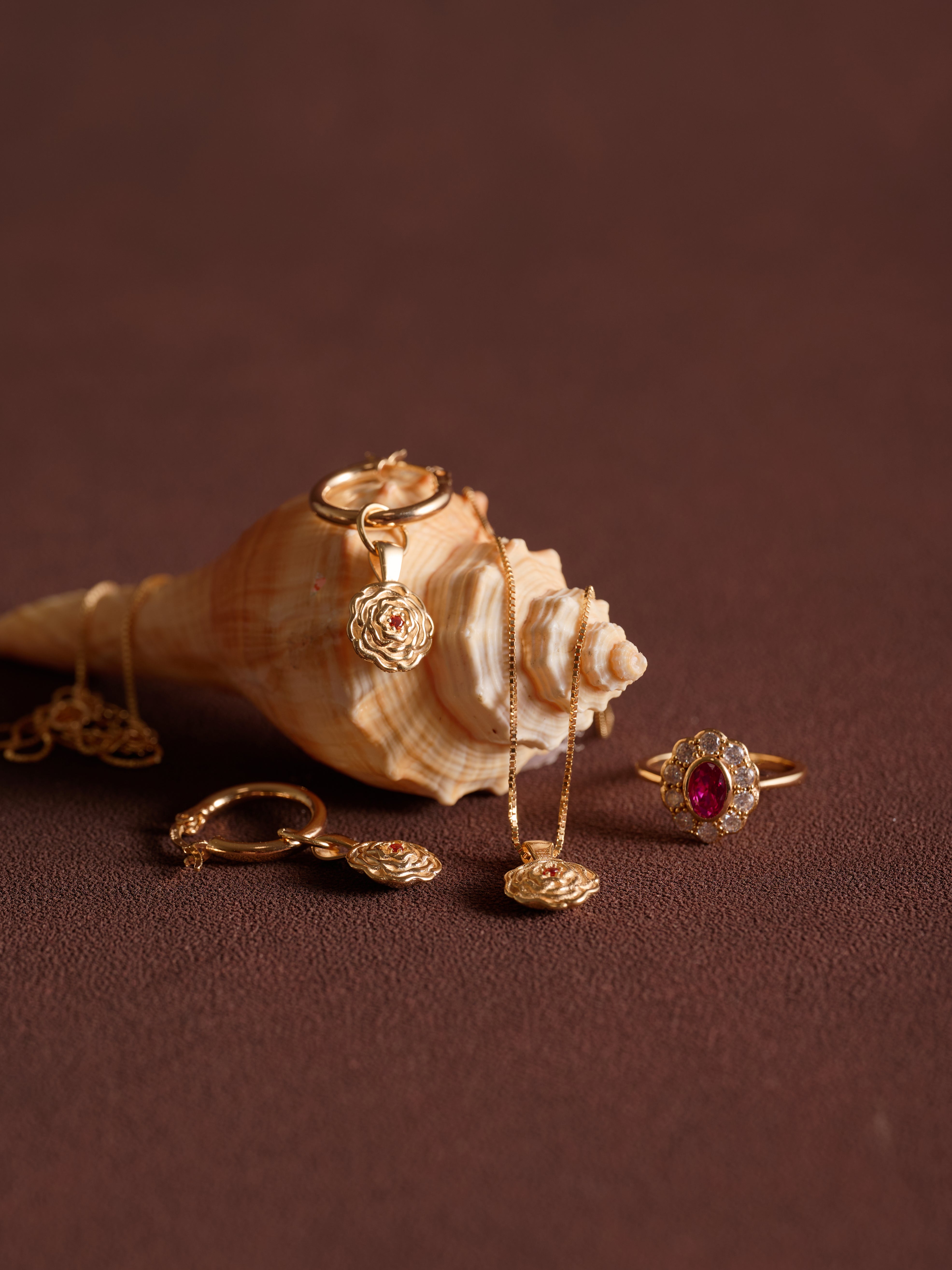 CAMELLIA’s Elsa Birth Month Necklaces are displayed with gold vermeil rings and a vintage floral pendant necklace beside a large seashell on a brown surface.