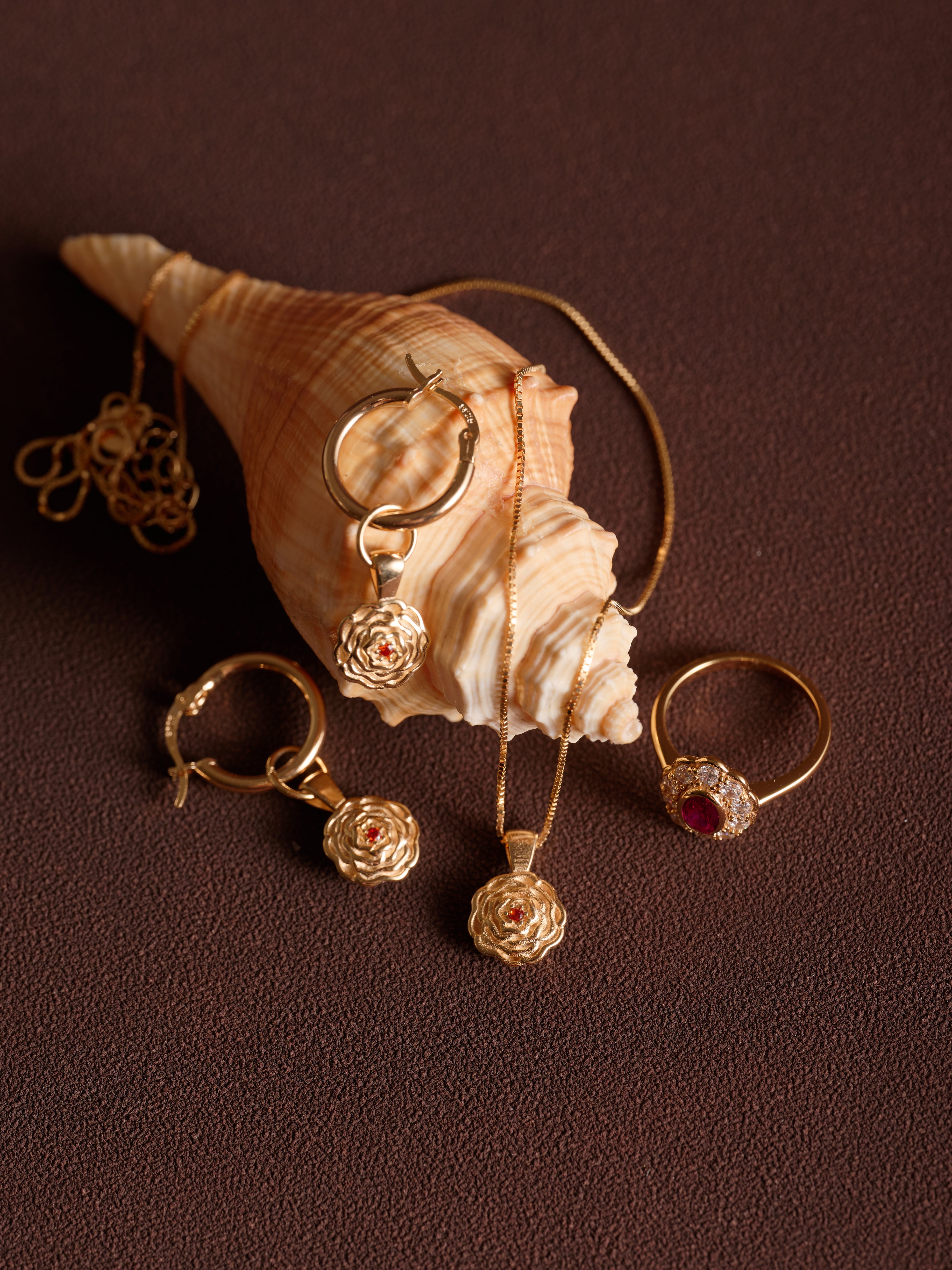 CAMELLIA Elsa Birth Month Necklaces, featuring vintage floral pendants and ruby accents, are artfully arranged around a large conch shell on a brown textured surface.