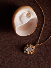 The CAMELLIA Capri Moonstone Necklace, featuring gold vermeil and a filigree design with white moonstone, rests beside a seashell on a textured brown surface, reflecting the vintage-inspired charm of this elegant piece.