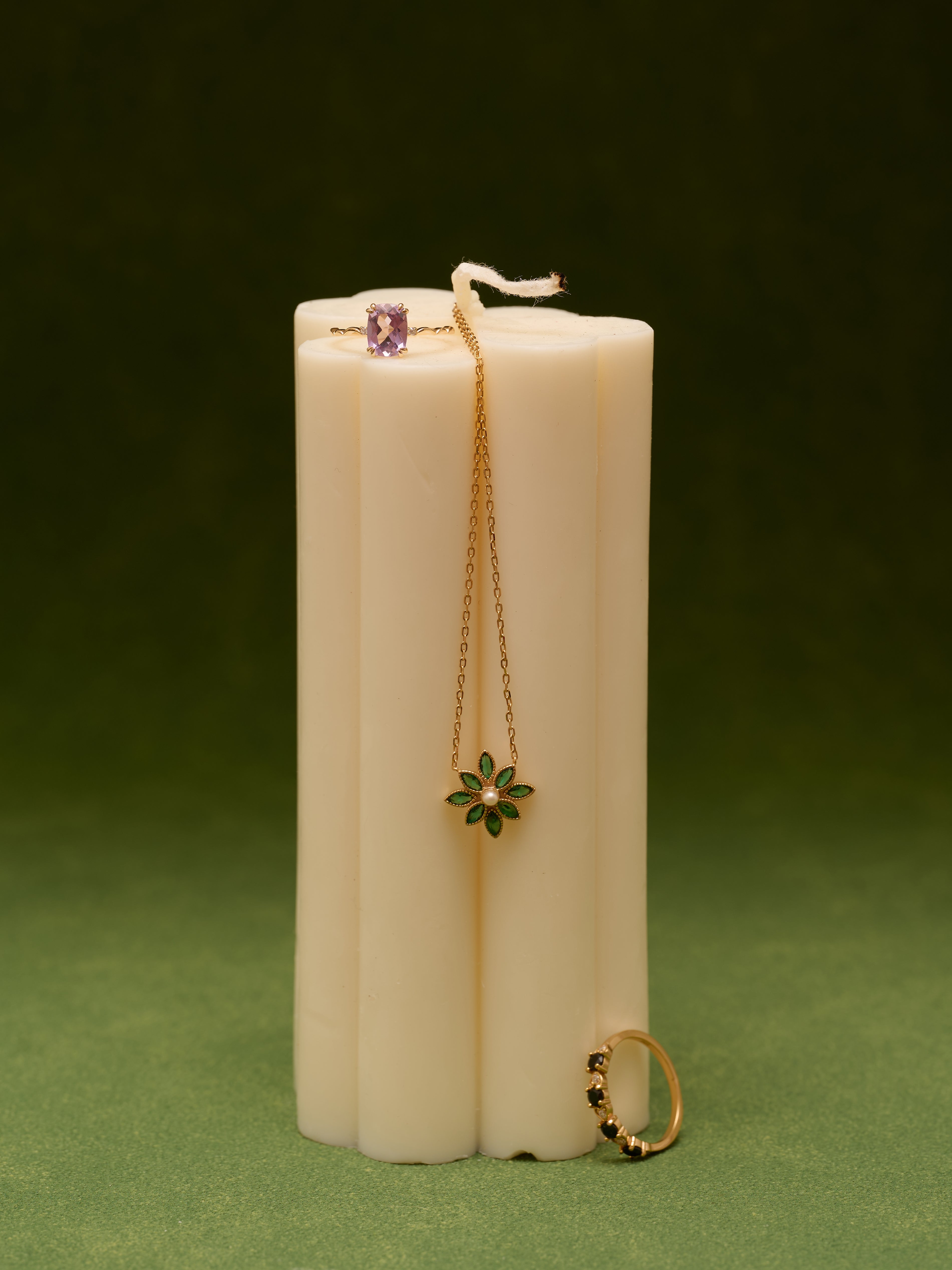 A white candle displayed vertically, adorned with a gold necklace featuring a green flower pendant, the CAMELLIA Giselle Onyx Ring, and a stackable ring topped with a purple stone.