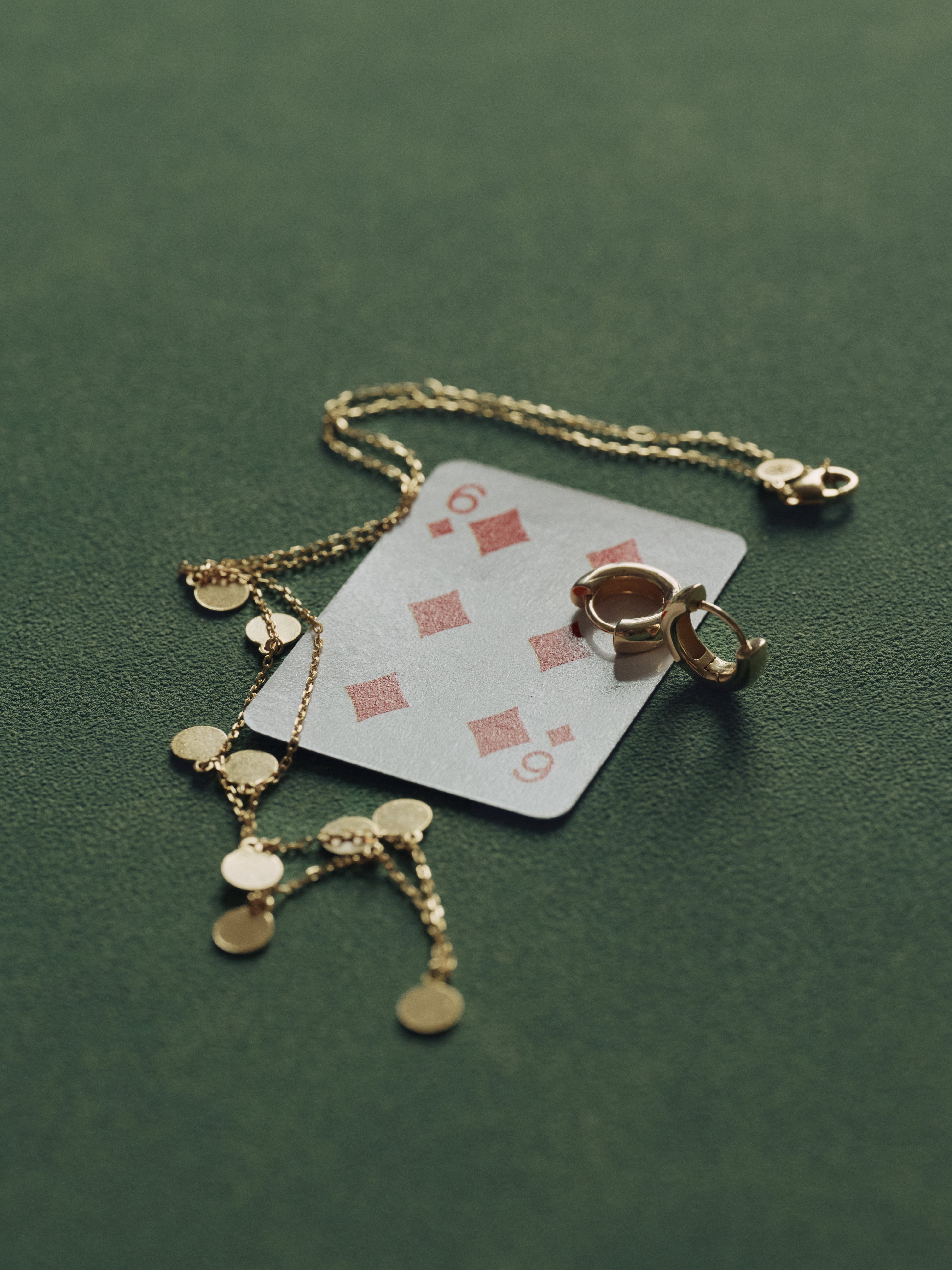 A six of diamonds playing card rests on a green surface, partially covered by a 14K gold vermeil chain necklace and CAMELLIA's hypoallergenic Mia Hoops earrings.