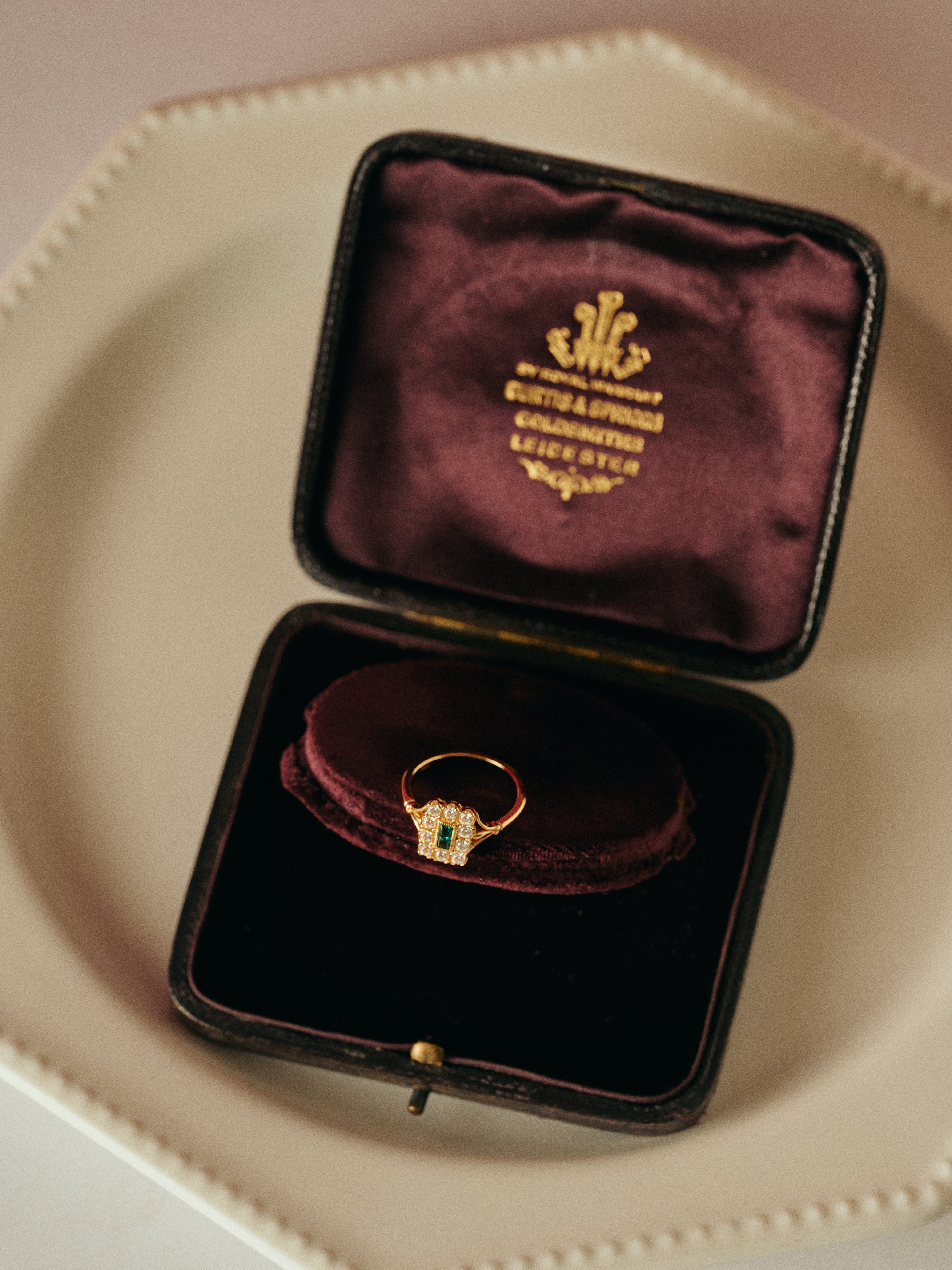 The CAMELLIA Seraphina Emerald Ring, featuring an emerald-green center, is showcased in a maroon velvet box with gold lettering on the lid atop a white plate, radiating timeless heirloom elegance.